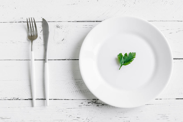 Table setting with white plate, modern cutlery (fork and knife) and little leaf of fresh parsley on white background, top view, copy space. Diet, fasting, spring, vegan raw food concept. - 写真・画像