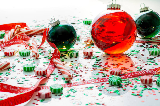 Διακόσμηση με το κόκκινο υγρό γεμάτο χριστουγεννιάτικο στολίδι μπάλα και δύο πράσινο στολίδι γεμάτο μπάλες, περιβάλλεται από μια κόκκινη κορδέλα έχουν τον εαυτό σας ένα καλά Χριστούγεννα, μια σειρά από χριστουγεννιάτικα λαμπάκια και μέντα καραμέλα - Φωτογραφία, εικόνα