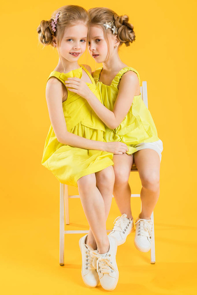 Twins girls are sitting on a chair on a yellow background. - Photo, Image