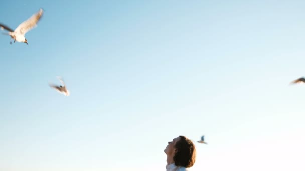 Europese jongeman is het voeden van meeuwen met brood aan de zee bij zonsondergang tegen een heldere blauwe hemel. Mooie man gooit voedsel voor zeevogels, een weergave van onderen - Video