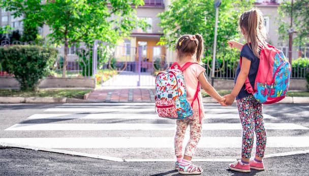 Los niños van a la escuela, los estudiantes felices con mochilas escolares y tomados de la mano, cruzan la calle, el concepto de educación con las niñas, el espacio para el texto
 - Foto, imagen