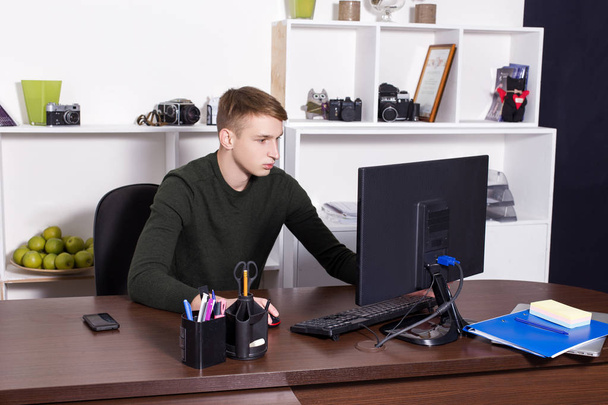A young guy dressed in a dark sweater works in the office at a table with a computer and talks on the phone. Business concept - Фото, изображение