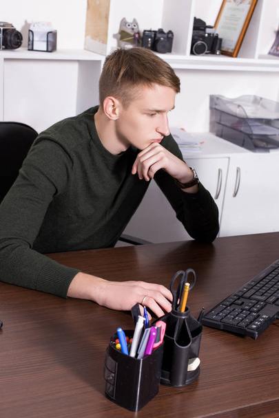 A young guy dressed in a dark sweater works in the office at a table with a computer and talks on the phone. Business concept - Φωτογραφία, εικόνα