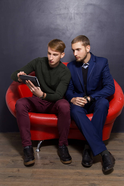 two guys in the official style are sitting with a tablet on a red sofa on a dark background. Business concept - Foto, imagen