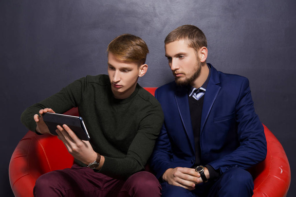Zwei Typen im offiziellen Stil sitzen mit einem Tablet auf einem roten Sofa vor dunklem Hintergrund. Geschäftskonzept - Foto, Bild