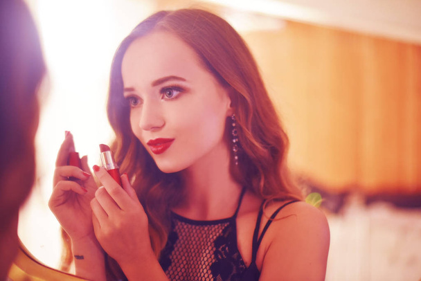 A beautiful young woman is doing herself a makeover. Girl with cerly hair doing evening makeup using lipstick in front of mirror in red light. Clouse up girl with red lips. - Foto, Imagen