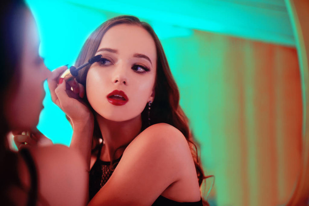 A beautiful young woman is doing herself a makeover. Girl with cerly hair doing evening makeup using Mascara in front of mirror in neon light. Clouse up girl with red lips. - Photo, image