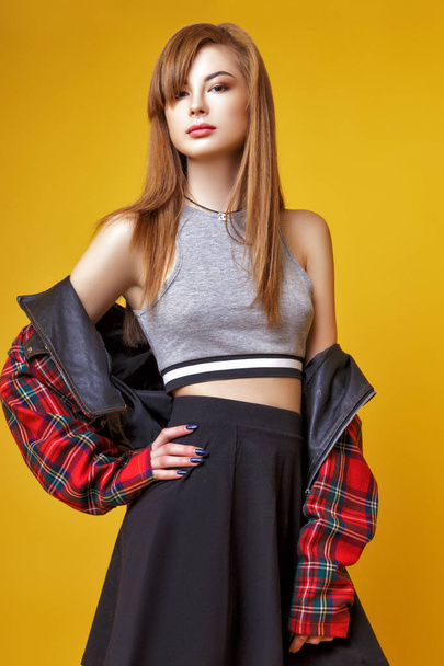 A cute young Asian girl with red hair, dressed in a plaid jacket, skirt and leggings posing in the studio on a yellow background. - Foto, Imagen