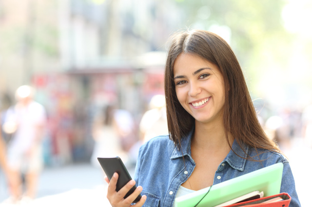 Happy studentka lodě, při pohledu na fotoaparát drží chytrý telefon a složky na ulici - Fotografie, Obrázek
