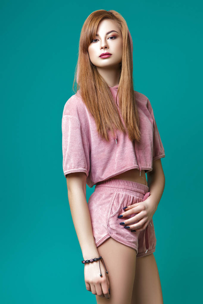 A cute young Asian girl with red hair, dressed in pink shorts and a sweatshirt with hood, posing in the studio on a green background. - Foto, Imagen