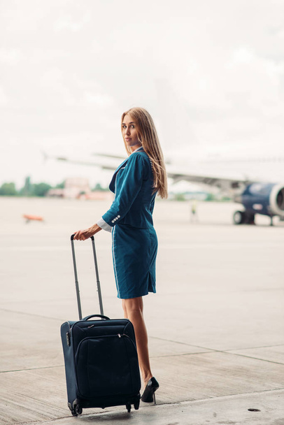 Young stewardess with suitcase on aircraft parking, tail of the airplane on background. Air hostess in suit with luggage - Фото, изображение