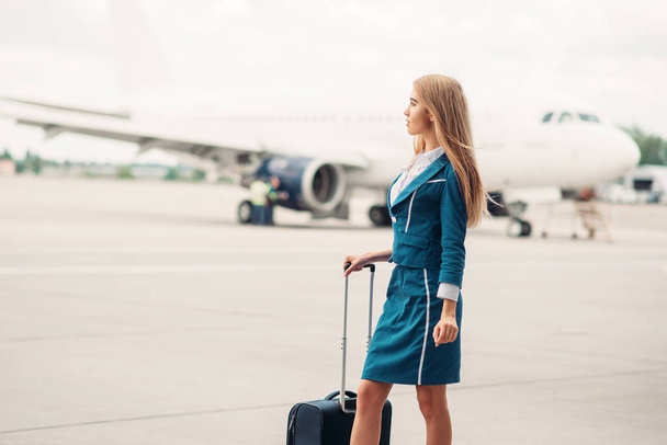 Сексуальная стюардесса с чемоданом на парковке самолетов, самолет на заднем плане
. - Фото, изображение