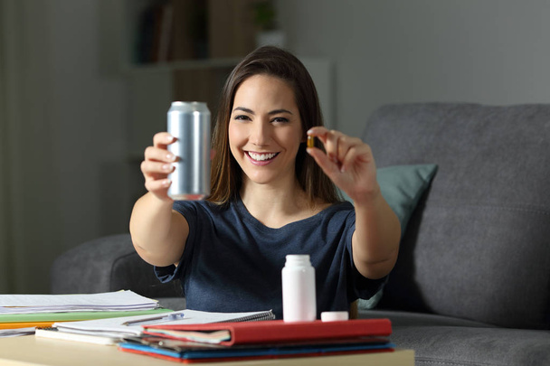Front view portrait of a happy student showing energy drink and vitamin pill in the night at home - Φωτογραφία, εικόνα