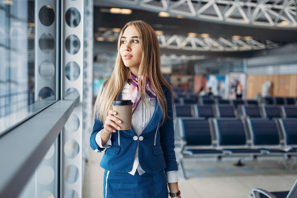 Stewardess with suitcase and coffee against window in airport. Air hostess with baggage in waiting room, flight attendant with hand luggage, aviatransportations job - Foto, afbeelding
