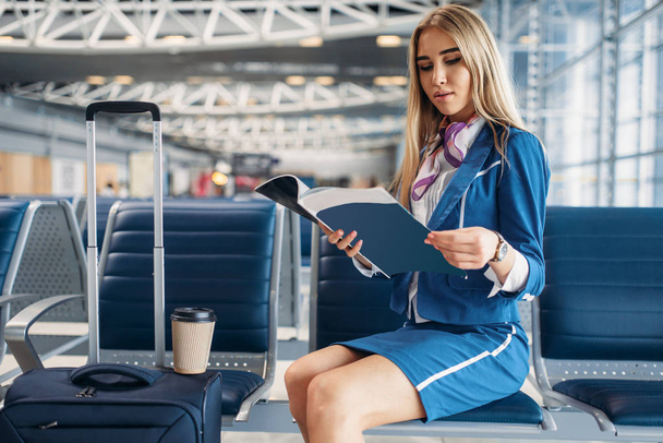 Αεροσυνοδός με βαλίτσα που κάθεται στο κάθισμα στο χώρο αναμονής στο αεροδρόμιο και διαβάζει περιοδικό. Air οικοδέσποινα με αποσκευές, αεροσυνοδός με χειραποσκευές, aviatransportations εργασία - Φωτογραφία, εικόνα