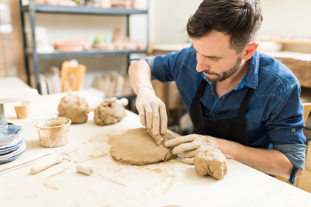 Artista masculino adulto medio que usa esponja para alisar la arcilla en la mesa en el taller de cerámica
 - Foto, imagen