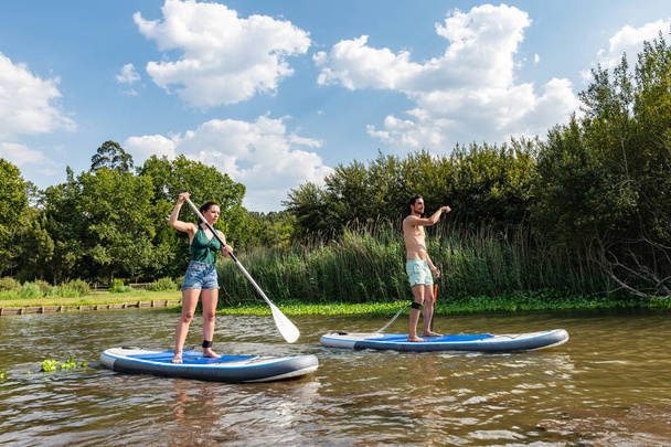 Man and woman stand up paddleboarding on lake. Young couple are doing watersport on lake. Male and female tourists during summer vacation. - Foto, imagen