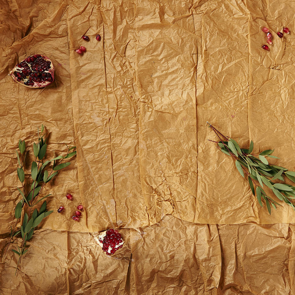 Crumpled Craft Paper Background with Pomegranate and Greens Top View. Wrinkled Brown Wrapping Paper Texture with Frame of Herbs and Red Garnet Seeds - Foto, Imagen
