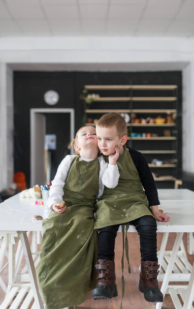Brother and little sister in aprons, sitting on a big white table in creative studio - Foto, Bild