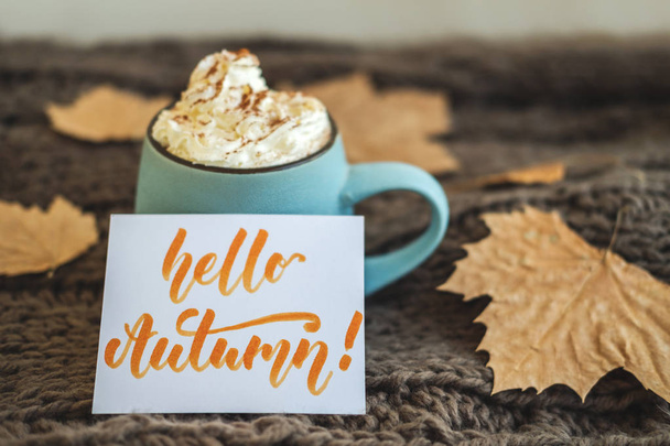 Mug of coffee, cocoa or hot chocolate with whipped cream and cinnamon on scarf with leaves, card Hello Autumn. Pumpkin latte - cozy drink for cold autumn or winter. - Foto, Bild