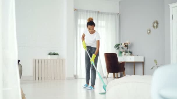 Pretty African American girl is mopping floor at home with flat plastic mop wearing gloves and wear-at-home clothing. Modern interiors, cleanness and people concept. - Imágenes, Vídeo