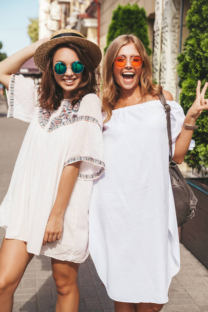 Fashion portrait of two young stylish hippie brunette and blond women models in summer sunny day in white hipster clothes posing on the street background. Going crazy - Fotografie, Obrázek