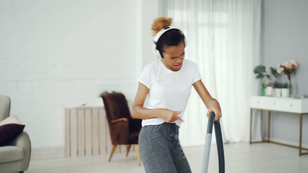 Active girl housewife is tidying apartment using vacuum cleaner and listening to music with headphones, dancing and singing. Joy, housework and youth concept. - Footage, Video