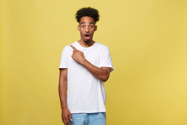 Astonished young African American man dressed in casual white shirt having excited fascinated look, pointing index finger at copy space on golden yellow background for your text or promotional content - Foto, afbeelding