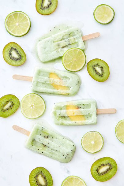 Summer pattern of frozen lime kiwi juice on a stick with a piece of peach and pieces of fruit on a gray background. Healthy dessert. Flat lay - Foto, Imagen