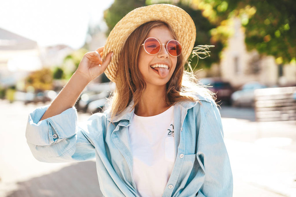 Portrait of beautiful cute smiling blond teenager model in summer hipster clothes posing on the street background. Showing her tongu - Zdjęcie, obraz