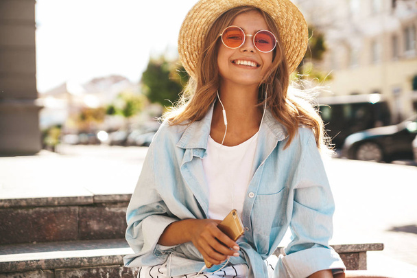 Portrait of beautiful cute smiling blond teenager model in summer hipster clothes sitting on the street background and listening music on her mobile phone - Fotoğraf, Görsel