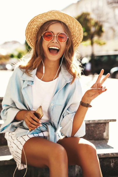 Portrait of beautiful cute smiling blond teenager model in summer hipster clothes sitting on stairs on the street background and listening music on her mobile phone. Showing peace sign, winking - Foto, immagini