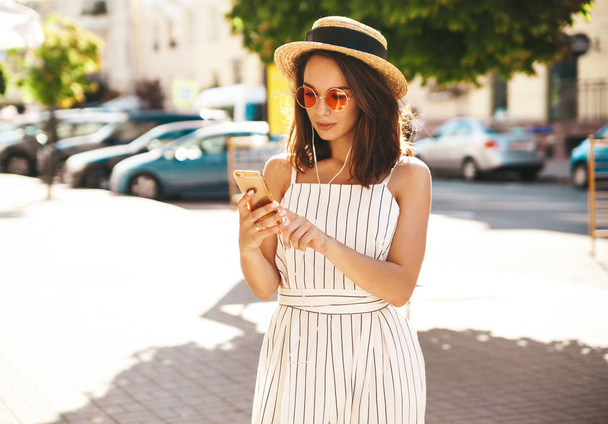 Porträt eines glücklichen Teenie-Mädchens in sommerlicher Hipster-Kleidung, das an einem sonnigen Tag auf der Straße Musik über Kopfhörer vom Smartphone hört - Foto, Bild