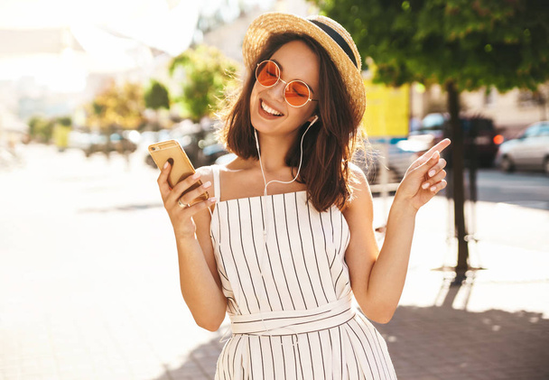 Portrait of happy smiling teen girl in summer hipster clothes listening music on her headphones from smartphone in the street on a sunny day - Фото, зображення