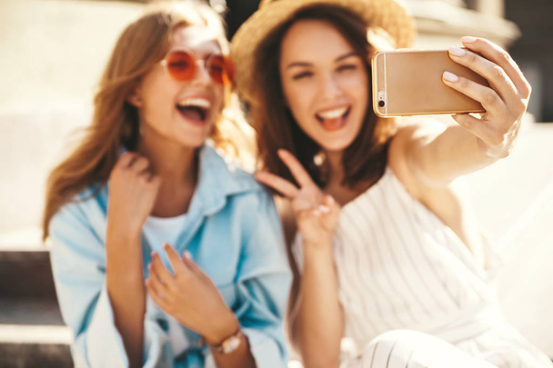 Two young female stylish hippie brunette and blond women models in summer sunny day in hipster clothes taking selfie photos for social media on smartphone on the street background. Showing peace sign - Foto, Imagen