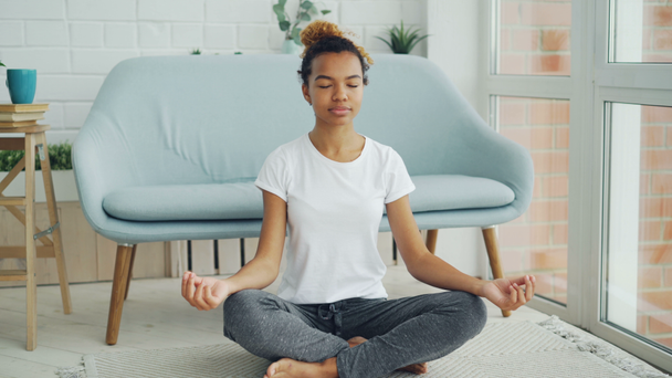 Vrij gemengd ras meisje zittend op de vloer in de lotuspositie met handen op knieën en mediteren is genieten van ontspanning en rust. Yoga, mensen en home concept. - Video