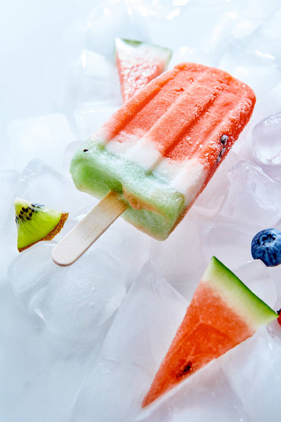 Ice lolly with vanilla smoothie on ice cubes and pieces of watermelon, blueberries, kiwi. Homemade cold healthy dessert. Top view - Fotoğraf, Görsel