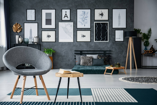 Grey armchair next to table on carpet in living room interior with gallery of posters. Real photo with blurred background - Foto, Imagem