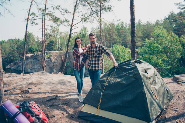 Green eco tourism. Young lovely people, couple wearing casual checkered shirts and jeans, standing together near set up tent in wild wood - Фото, зображення