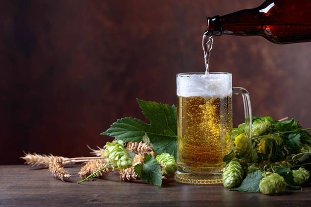 Ein Becher Bier, Getreide und Hopfen auf einem alten Holztisch. Bier wird aus der Flasche in einen Becher gegossen. Freiraum für Text. - Foto, Bild