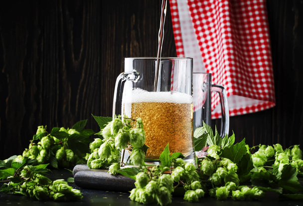 Large mugs with german light beer and fresh hops, pours into glass, dark table, selective focus - Fotografie, Obrázek