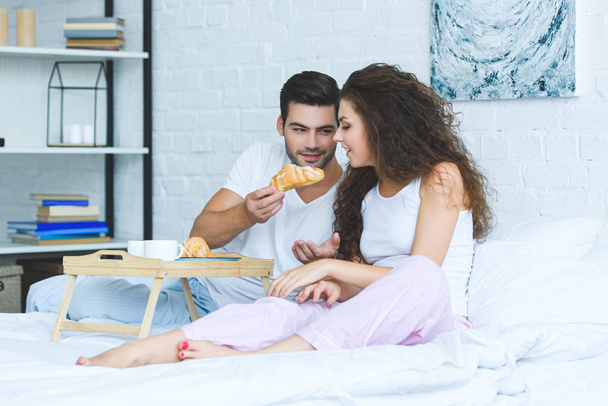 handsome young man feeding beautiful smiling girlfriend with croissant in bedroom - Zdjęcie, obraz