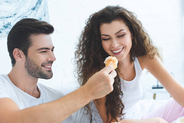 smiling young man feeding happy girlfriend with croissant for breakfast in bedroom - Фото, изображение