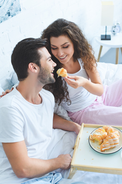 happy young woman feeding smiling boyfriend with croissant in bed  - 写真・画像