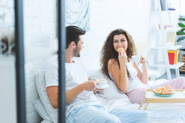 selective focus of happy young couple in pajamas having breakfast and smiling each other in bedroom  - Photo, Image