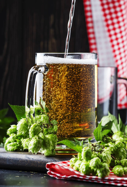 Large mugs with german light beer and fresh hops, pours into glass, dark table, selective focus - Photo, Image