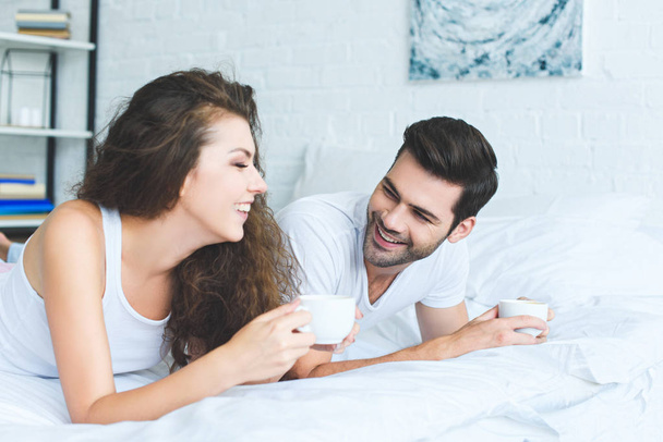 cheerful young couple in pajamas holding cups of coffee and lying on bed - Foto, Imagen