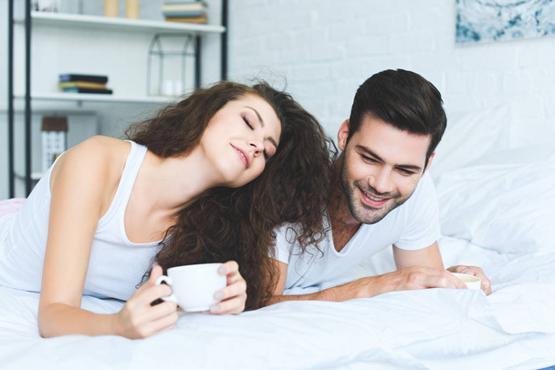 コーヒーのカップを保持していると、ベッドに横になっている若いカップルの笑顔が美しい  - 写真・画像