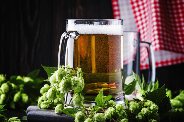 Large mugs with german light beer and fresh hops, pours into glass, dark table, selective focus - 写真・画像