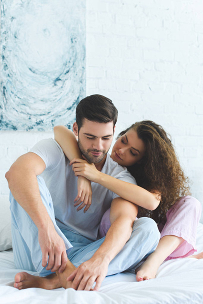 beautiful young woman hugging handsome husband while sitting together on bed - Photo, Image